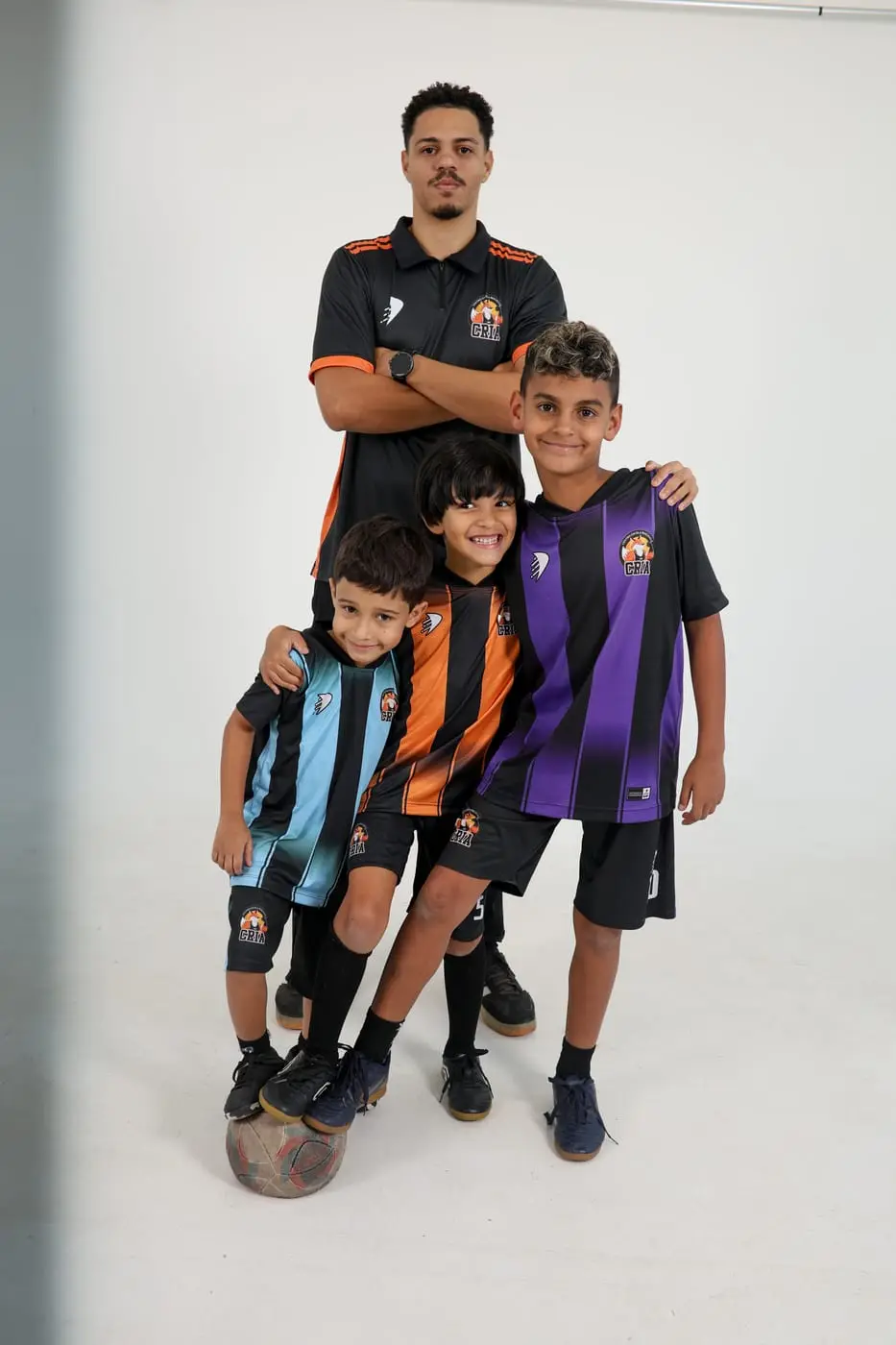 Três alunos pequenos com camisas de futebol e a bola, ao lado do professor